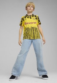 Camiseta de fútbol a rayas amarillas y negras con logo y nombre de la marca, combinada con jeans de pierna ancha en azul claro y zapatillas negras.
