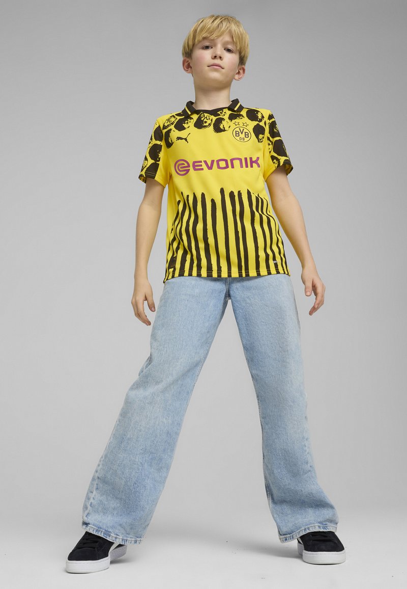 Camiseta de fútbol a rayas amarillas y negras con logo y nombre de la marca, combinada con jeans de pierna ancha en azul claro y zapatillas negras.