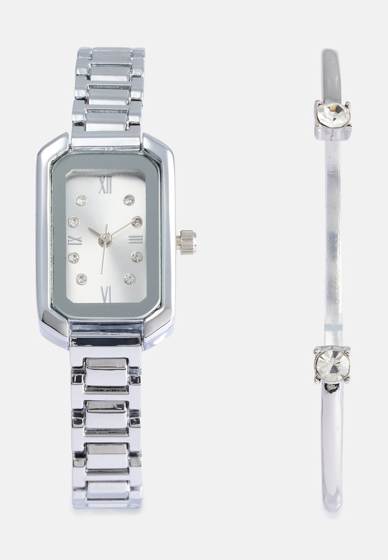 Relógio de pulso em metal prateado com mostrador rectangular, números romanos e detalhes em cristal; apresenta uma pulseira ligada e uma coroa para ajustes.