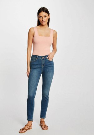 Donna con capelli lunghi e scuri che indossa una canottiera rosa chiaro, jeans skinny blu a vita alta e sandali marroni con cinturini, in piedi.