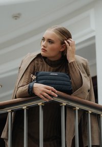 Zwarte puffer tas met een logo, vastgehouden door een persoon in een bruine geribde jurk en een bruine geblokte blazer, rustend op een houten leuning.