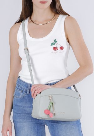 Žena v bílém tílku s výšivkou třešní a světle modrých džínách, držící světle šedou kabelku přes rameno s ozdobou ve tvaru třešní.