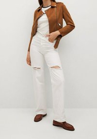 Giacca da biker in suede marrone, jeans bianchi strappati con buchi alle ginocchia e mocassini marroni. Linee pulite e colori a contrasto.