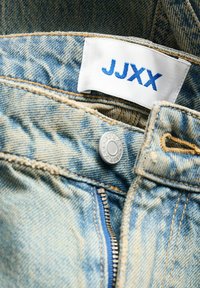 Light blue denim jeans with visible stitching, metal button closure, and a white label displaying "JJXX" in blue. Zipper detail included.