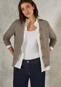 Šedý cardigan s ribbenou texturou, se pěti knoflíky a zkrácenými rukávy. Nosený přes bílou košili a spárovaný s tmavými džíny.