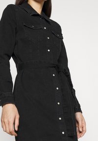 Vestido de mezclilla negra con manga larga, cierre de botones en el frente, dos bolsillos en el pecho, un cuello y una cintura con cinturón a juego.