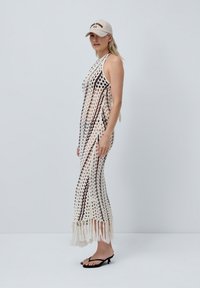 Robe en crochet crème avec des rayures verticales marron, longue jusqu'au sol, avec un ourlet à franges. Le mannequin porte une casquette beige et des tongs noires.