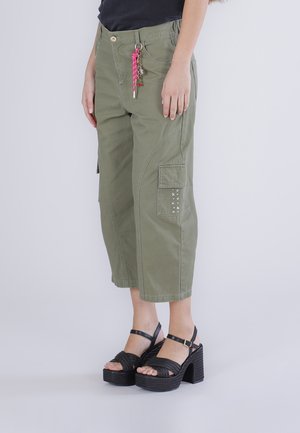 Persoon draagt olijfgroene cropped cargobroek met een roze sleutelhanger en zwarte platformsandalen.