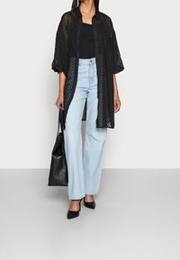 Sort, gennemsigtig skjorte med knapper og et struktureret mønster, parret med lyseblå bredbenede jeans og sorte spidse hæle. Sort håndtaske.