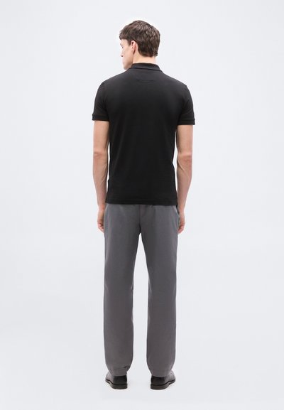 Homme debout, de dos, portant un polo noir à manches courtes, un pantalon gris et des chaussures noires, sur un fond clair uni.