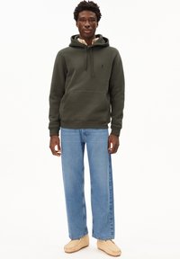 Felpa in pile verde scuro con tasca a marsupio, abbinata a jeans a gamba larga blu chiaro e scarpe slip-on beige.