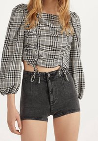 Žena v černých džínových šortkách s vysokým pasem a černobílém kostkovaném crop topu s nařasenými dlouhými rukávy.