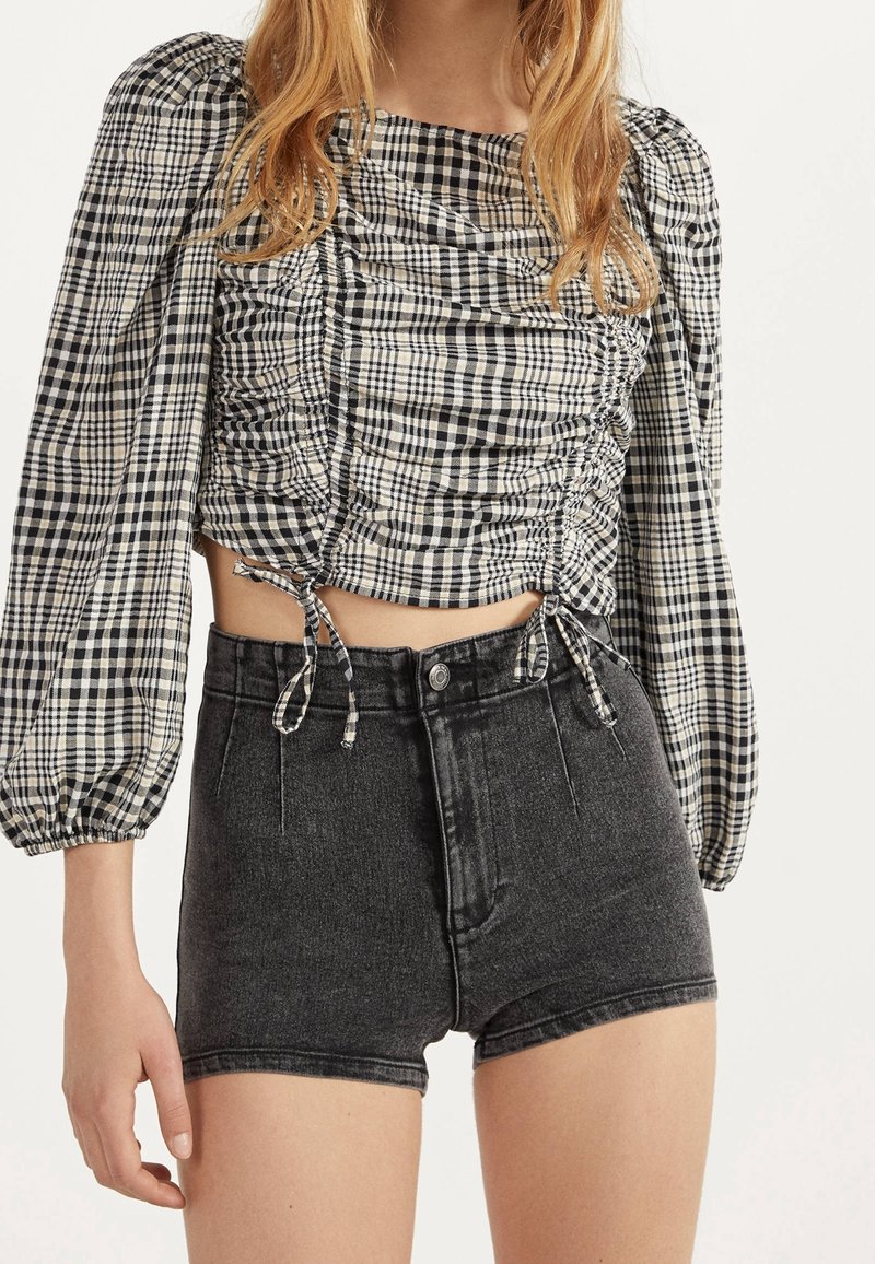 Žena v černých džínových šortkách s vysokým pasem a černobílém kostkovaném crop topu s nařasenými dlouhými rukávy.