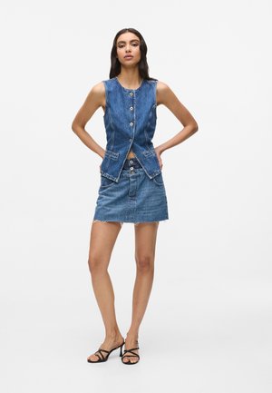 Γυναίκα με αμάνικο τζιν γιλέκο με κουμπιά, συνδυασμένο με τζιν φούστα ψηλής μέσης και μαύρα σανδάλια με λουράκια και τακούνι, στέκεται με τα χέρια στη μέση.