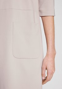 Ärmlös, ljusbeige klänning med en mjuk textur, som har en stor, rund ficka framtill och en söm över bröstområdet.