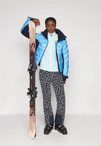 Kék puffert dzseki, világoskék alapréteg, fekete-fehér mintás sí nadrág, fekete kötésekkel ellátott sílécet tart, fekete sícipő.