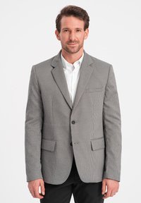 Grauer, strukturierter Blazer mit kariertem Muster, Ein-Knopf-Verschluss, Reverskragen und zwei Fronttaschen, getragen über einem weißen Hemd.