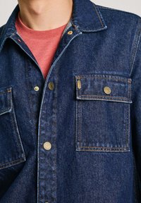Giacca di jeans blu con due tasche sul petto con bottone, dettagli in hardware dorato e cuciture in contrasto color sabbia. Indossata sopra una camicia rossa.