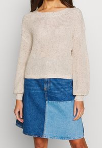 Woman wearing a beige knit sweater with balloon sleeves and a blue patchwork denim skirt, standing against a plain background.