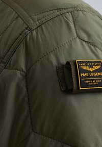 Grüne gepolsterte Jacke mit diagonalen Nähten, ausgestattet mit einem schwarzen und gelben Etikett mit der Aufschrift "PME LEGEND", befestigt mit einem Stoffband.
