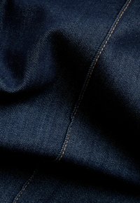 Tumma farkkukangas, jossa on sileä pinta ja näkyvät ompeleet kontrastivärillä oranssina. Pinnassa on hieno kiilto.