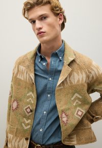 Giacca marrone con motivo geometrico e tonalità terrose, indossata sopra una camicia di jeans blu con colletto classico e chiusura a bottoni.