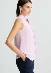 Lichtroze mouwloze blouse met een strikdetail bij de kraag, met een soepele textuur en een losse pasvorm, gecombineerd met marineblauwe broek.