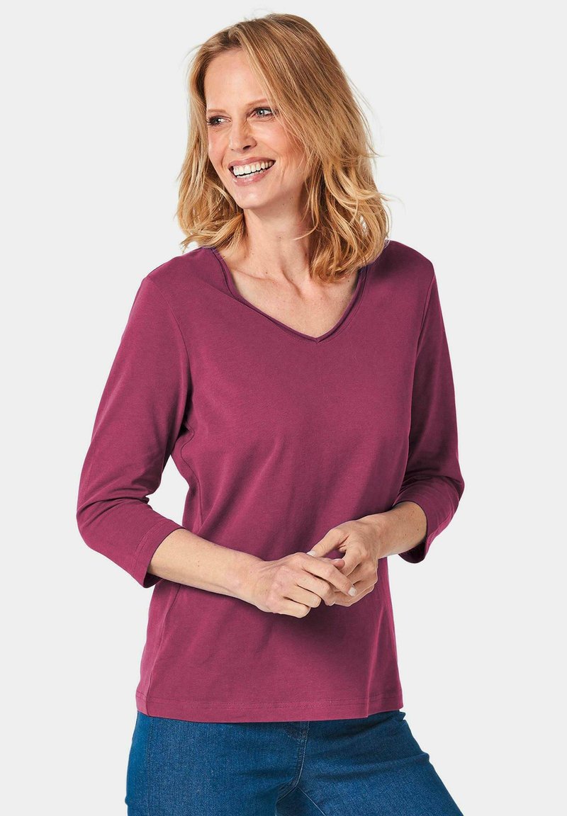 V-Ausschnitt, Bluse mit drei Viertelärmeln in tiefem Pflaumenton, aus weichem Stoff gefertigt. Sie weist eine lockere Passform und eine einfache Naht entlang des Ausschnitts auf.