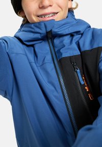 Enfant souriant portant un appareil dentaire et une veste d'hiver bleue avec une poche poitrine noire et un logo orange, zipée jusqu'au cou.