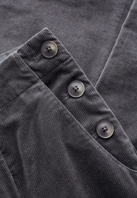 Pilkas aksominio audinio, kuriame trys tamsios mygtukai ir struktūrizuotas dizainas su vertikaliomis briaunomis. Kraštai tvarkingai apdirbti.