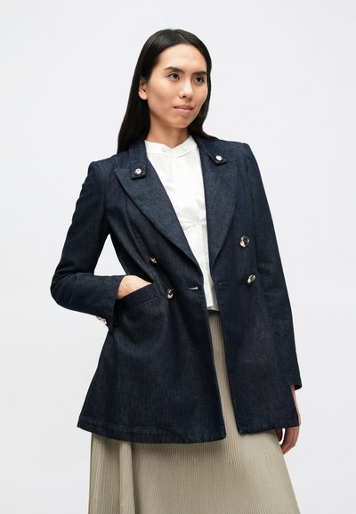 Σκούρο τζιν σακάκι διπλής επένδυσης με χρυσαφί κουμπιά, γιακάδες με εγκοπές και πλαϊνές τσέπες, φορεμένο πάνω από λευκό πουκάμισο και πλισέ φούστα.