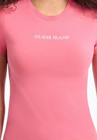Rožinė medvilninė marškinė su trumpomis rankovėmis ir apvalia iškirpte; krūtinėje turi baltą "GUESS JEANS" logotipą.
