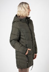Frau mit kurzen blonden Haaren, die eine olivgrüne, gesteppte Winterjacke mit Kapuze trägt, mit den Händen in den Reißverschlusstaschen, die nach rechts blickt, vor einem weißen Hintergrund.