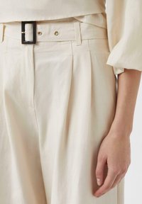 Kremowe spodnie z zakładkami, szerokim pasa, oraz czarną prostokątną klamrą. Tkanina ma gładki, teksturowany wygląd.