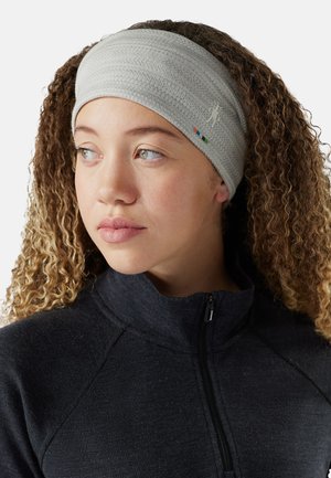 Ung person med krøllet hår iført et gråt tekstureret pandebånd og en mørk zip-up jakke, der ser lidt til venstre mod en hvid baggrund.