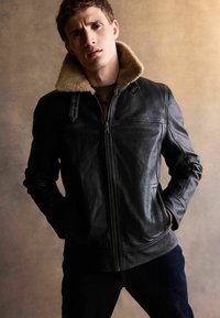 Jachetă din piele neagră cu guler din blană de oaie, închidere cu fermoar, buzunare laterale și accente minime din metal. Suprafață texturată cu un design ajustat.