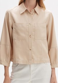 Beige cropped shirt met een knoopsluiting aan de voorkant, twee borstzakken en 3/4 mouwen. Glad materiaal met zichtbare stikseldetails.
