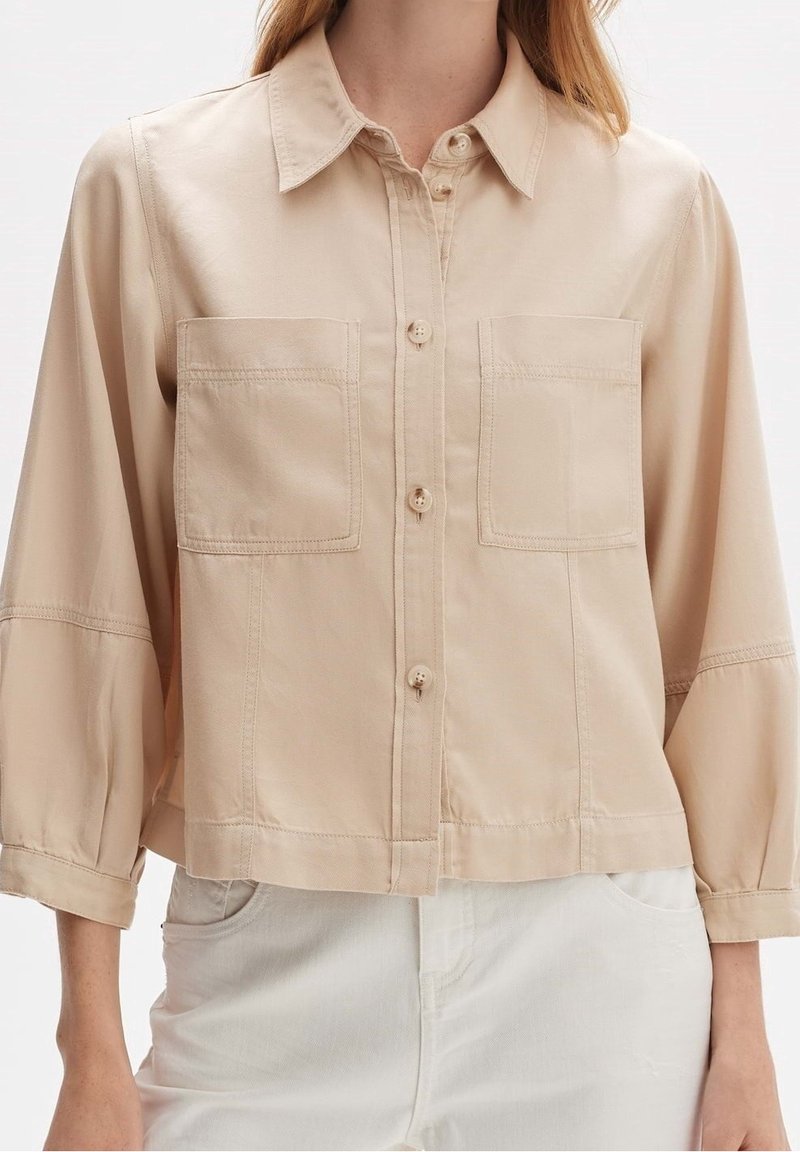 Beige cropped shirt met een knoopsluiting aan de voorkant, twee borstzakken en 3/4 mouwen. Glad materiaal met zichtbare stikseldetails.