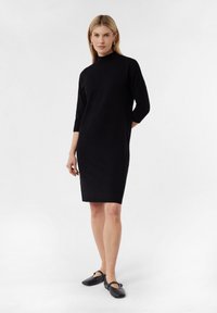 Schwarzes Strickkleid mit hohem Ausschnitt, Dreiviertelärmeln und gerader, knielanger Schnitt; kombiniert mit schwarzen flachen Schuhen.