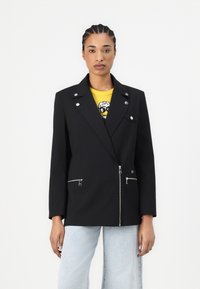 Blazer nero con bottoni e zip argentati, vestibilità sartoriale, spalle strutturate, maglione giallo a contrasto sotto, e pantaloni in denim chiaro.
