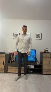 Un homme en chemise blanche et pantalon sombre se tient pieds nus dans un salon avec une télévision, des meubles en bois, des photos encadrées et des objets décoratifs.