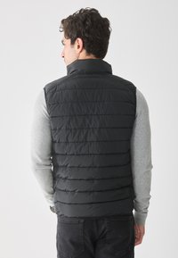 Chaleco de plumas negro con diseño acolchado horizontal, cuello alto y cierre con cremallera. Lleva puesto una camiseta de manga larga gris claro.