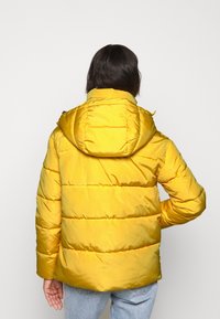 Gele gewatteerde jas met capuchon, voorzien van horizontale stiknaden en een gladde textuur. Achteraanzicht toont een ontspannen pasvorm en casual stijl.