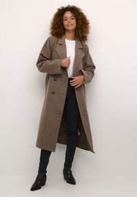 Brązowy wełniany płaszcz dwurzędowy z szerokimi klapami, w oversize'owym fasonie, z dwoma dużymi guzikami. Stylizowany z białym T-shirtem i czarnymi spodniami.