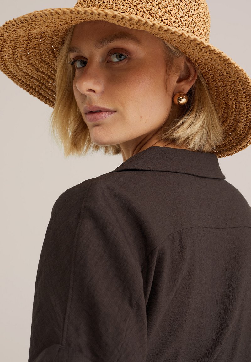 Femme blonde portant un chapeau de paille à large bord, des boucles d'oreilles en or et une chemise texturée marron foncé, regardant par-dessus son épaule.