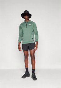 Πράσινο φλις με φερμουάρ, μακριά μανίκια και πλαϊνή τσέπη, συνδυασμένο με μαύρο σορτς. Φορεμένο με μαύρο καπέλο τύπου bucket και γυαλιά ηλίου. Μαύρα αθλητικά παπούτσια.