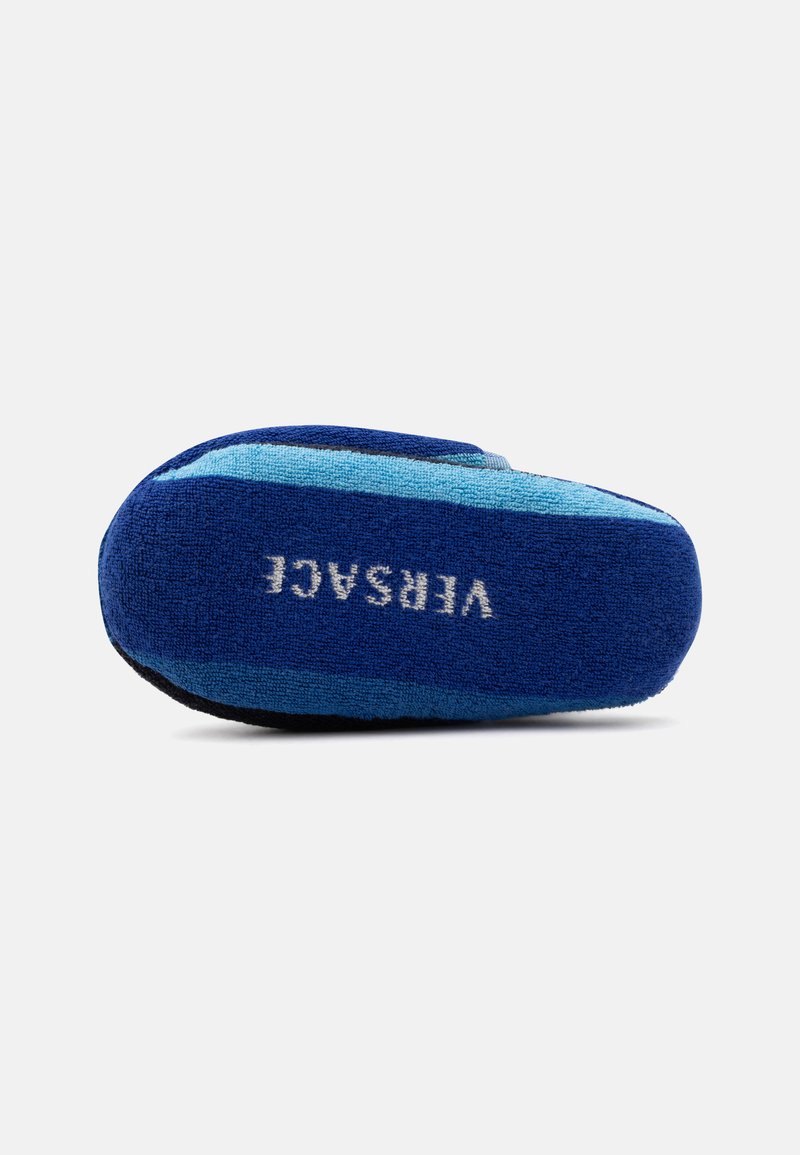 Blaue Hausschuhe mit hellblauen Streifen und dem Wort „VERSACE“, weiß auf der Sohle gestickt, von unten gezeigt.