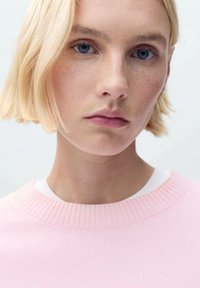 Różowy sweter z ściągaczem, nałożony na biały t-shirt typu crew neck. Teksturowana tkanina i miękkie wykończenie. Modelka ma krótkie blond włosy.