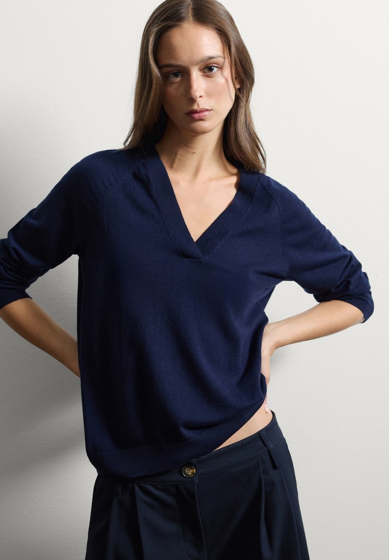 Maglione lavorato a maglia blu navy con scollo a V, maniche a tre quarti e una texture liscia. Indossato con pantaloni neri a vita alta, plissettati.