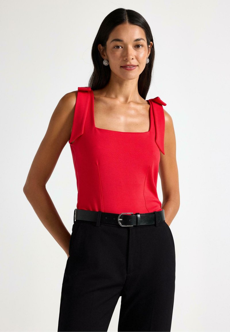 Červený top bez rukávů se čtvercovým výstřihem a mašličkovými detaily na ramínkách, nošený s černými kalhotami s vysokým pasem a černým opaskem.